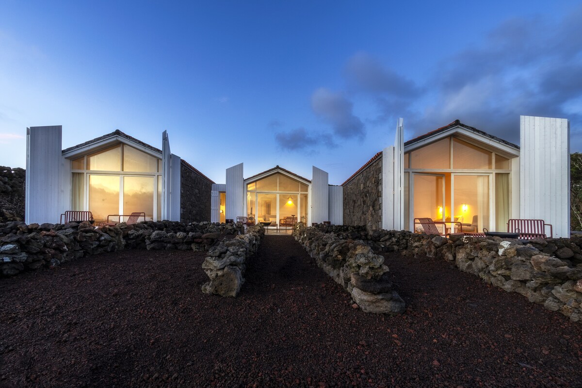 Three modern structures with large glass doors are positioned symmetrically, framed by natural stone pathways. Each building features welcoming lighting that enhances the tranquil ambiance. The surrounding landscape showcases volcanic rock, creating a harmonious blend with the architecture.