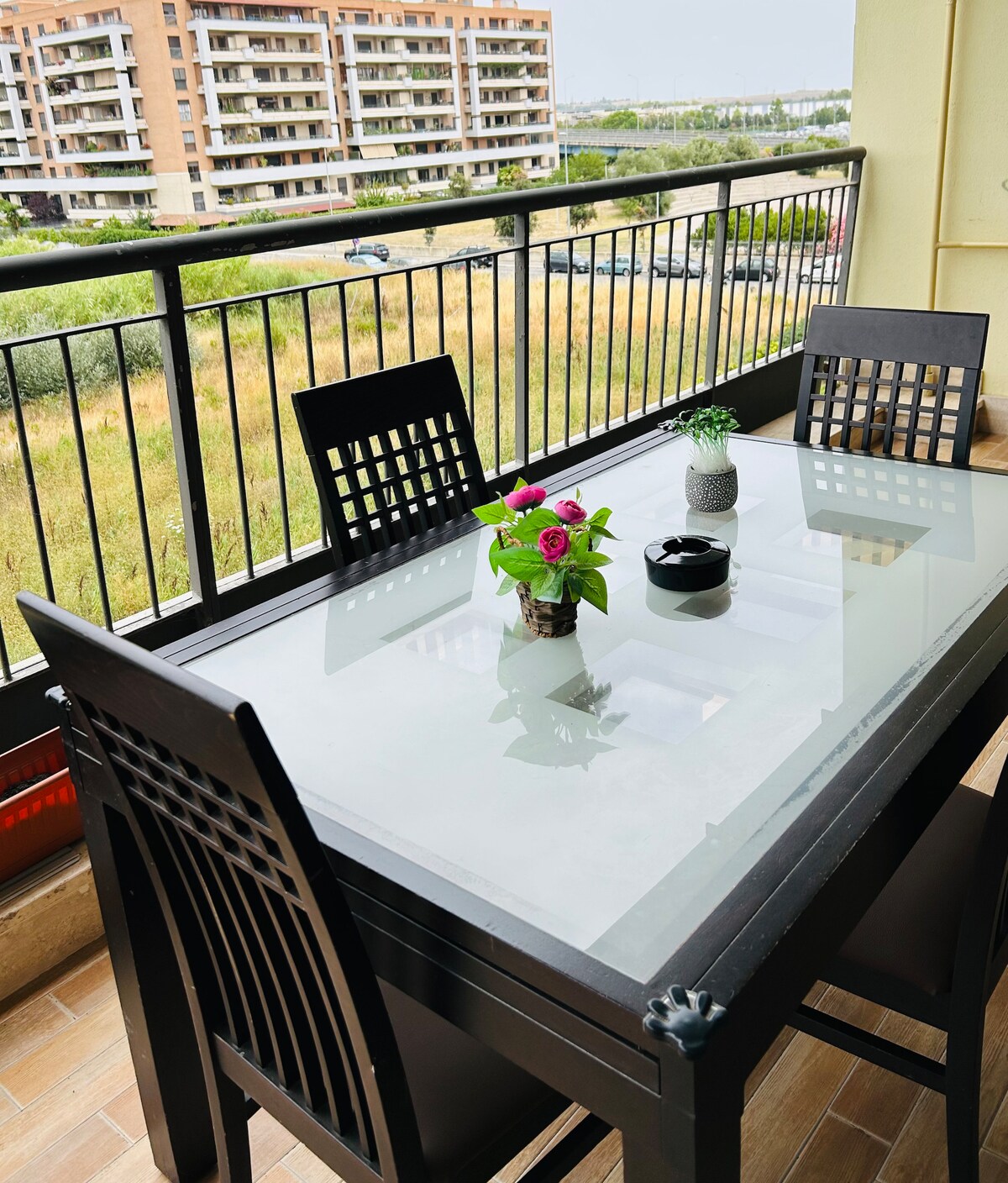 A balcony dining area is featured, showcasing a glass-top table surrounded by four black chairs. A small potted plant is positioned at the center, with additional decorative items off to the side. A view of surrounding greenery and a building is visible in the background.
