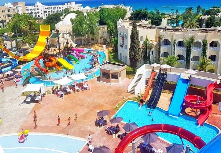 A vibrant outdoor water park showcases colorful slides and pools, with families enjoying the space. Umbrellas provide shade over seating areas, while lush greenery surrounds the facilities. The sparkling water reflects sunlight, creating a lively atmosphere.