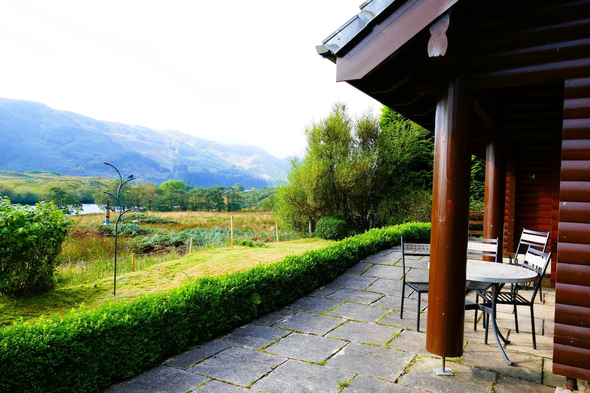 A patio area is visible, featuring a round table surrounded by several chairs. The space is bordered by neatly trimmed hedges, with views of the surrounding hills and lush greenery. The log house structure provides a rustic touch to the tranquil outdoor setting.