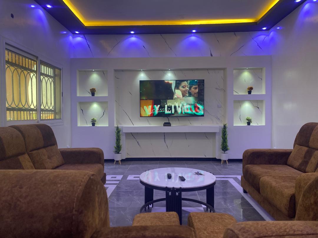 A modern living room features a comfortable seating area with plush sofas arranged around a circular coffee table. A large television is mounted on a sleek wall, flanked by illuminated shelves displaying decorative plants. Soft lighting enhances the contemporary design.