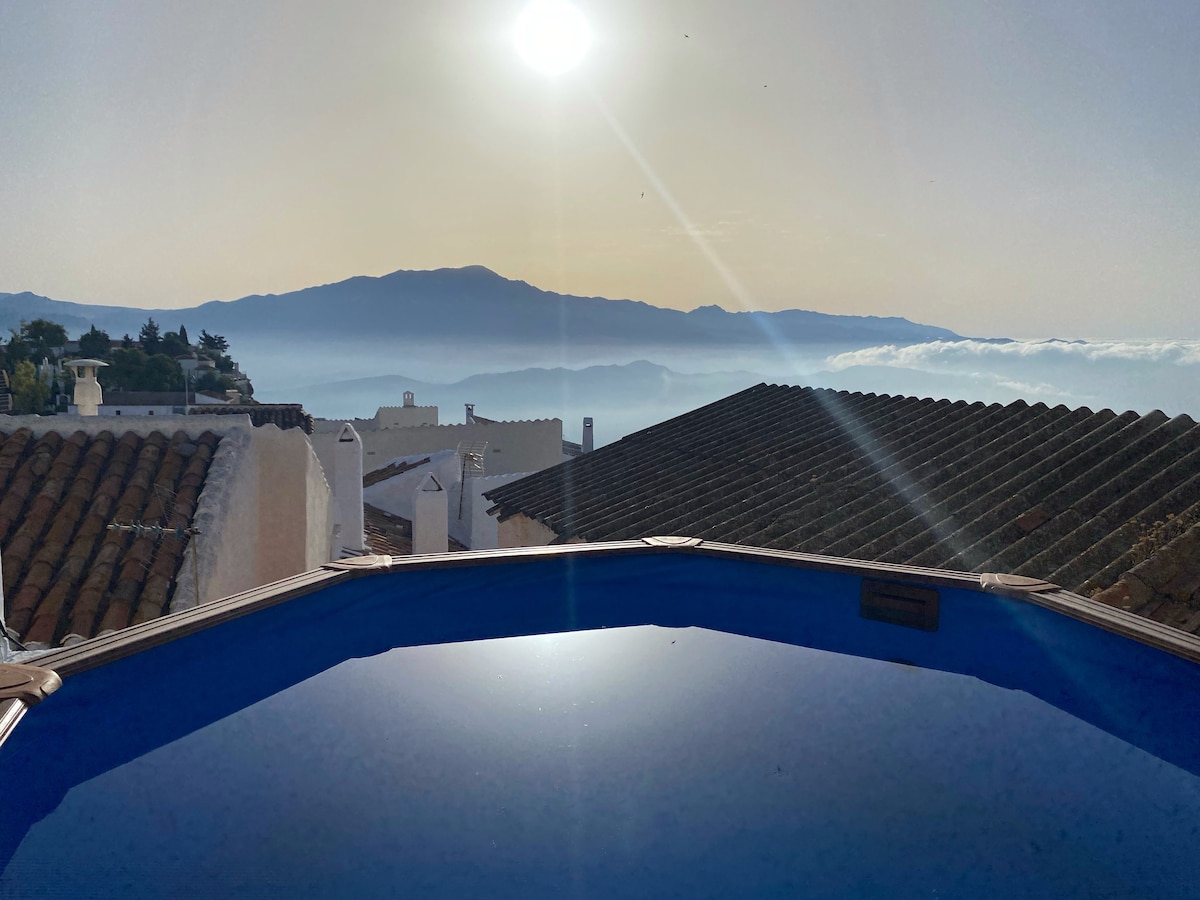A view from the dip pool showcases a serene landscape, with rooftops in the foreground and distant mountains rising above the sea of clouds. The sun casts a gentle glow on the water surface, creating an inviting atmosphere.
