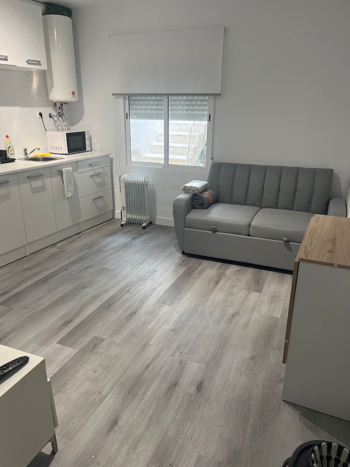 A small living area features a light grey couch with a rolled blanket, positioned near a window that allows natural light to fill the space. A compact kitchen with modern cabinetry and appliances is visible in the background, alongside a small wooden table.