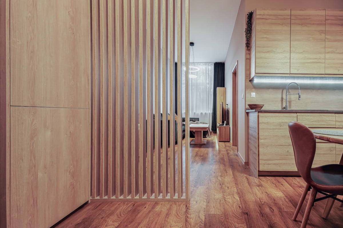 A modern open interior features wooden flooring and light wood cabinetry. A wooden slat partition provides subtle separation between living spaces. Soft lighting illuminates the room, highlighting a kitchen area, dining table, and a cozy seating arrangement visible in the background.