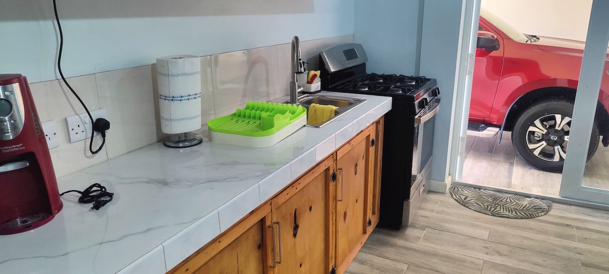 A well-equipped kitchen features a marble countertop with wooden cabinetry. A sink, dish drying rack, and a coffee maker are visible. The kitchen includes a modern stove and has a window leading to an outdoor space, revealing part of a parked vehicle.