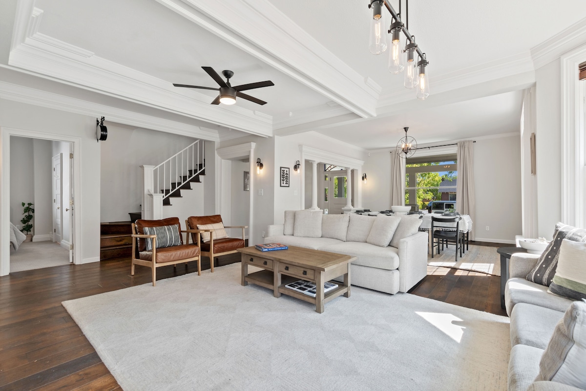 The open-concept living area features a spacious light-filled interior. A large sectional sofa faces a wooden coffee table, while stylish armchairs add to the seating. Natural light streams through large windows, highlighting the elegant ceiling details and wooden flooring.