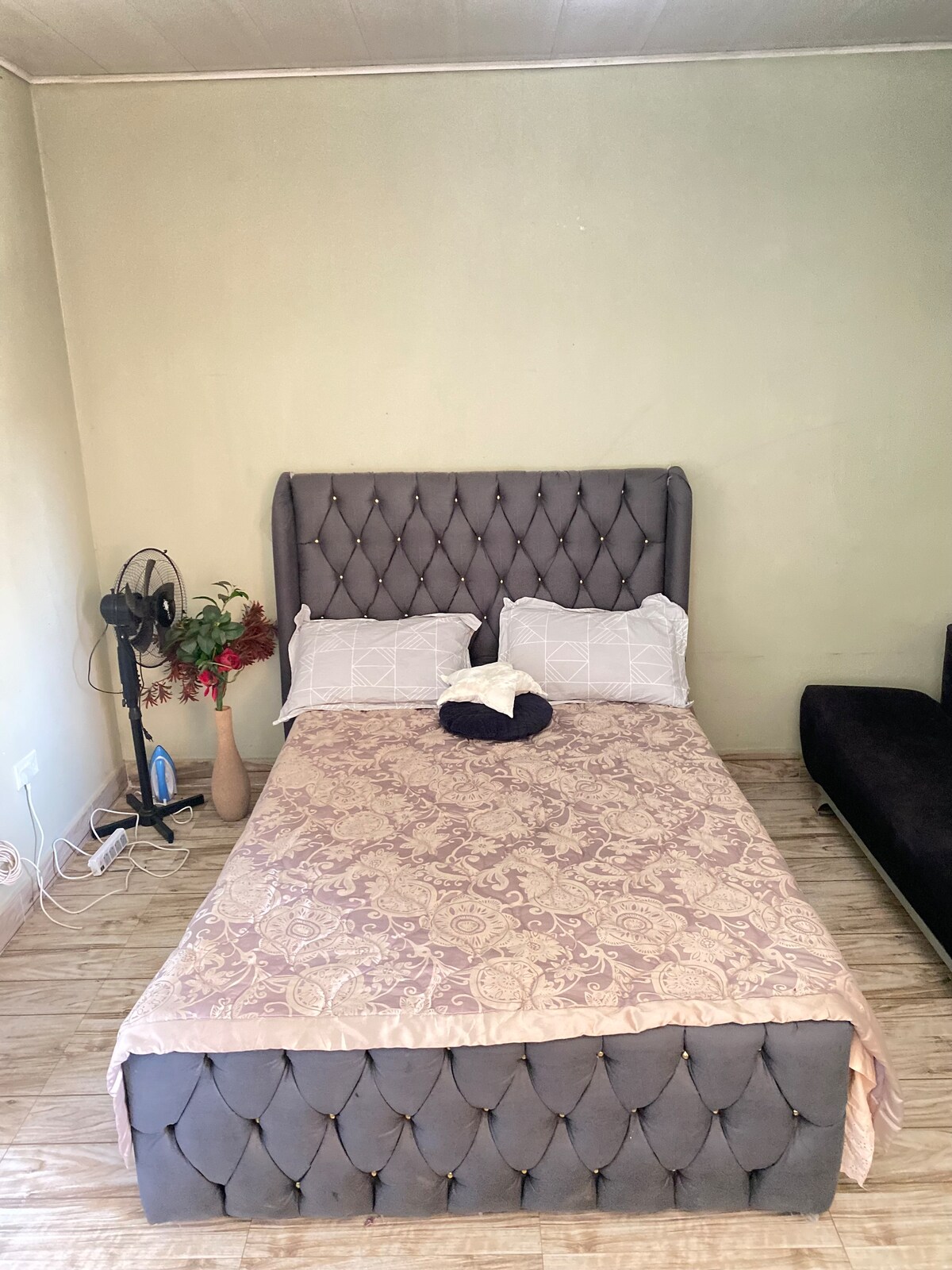 A spacious bed with a tufted grey headboard is dressed in a light-colored patterned duvet. Two pillows rest at the head of the bed. A small fan and decorative vase with flowers are positioned beside the bed, while a dark seating area is visible in the background.