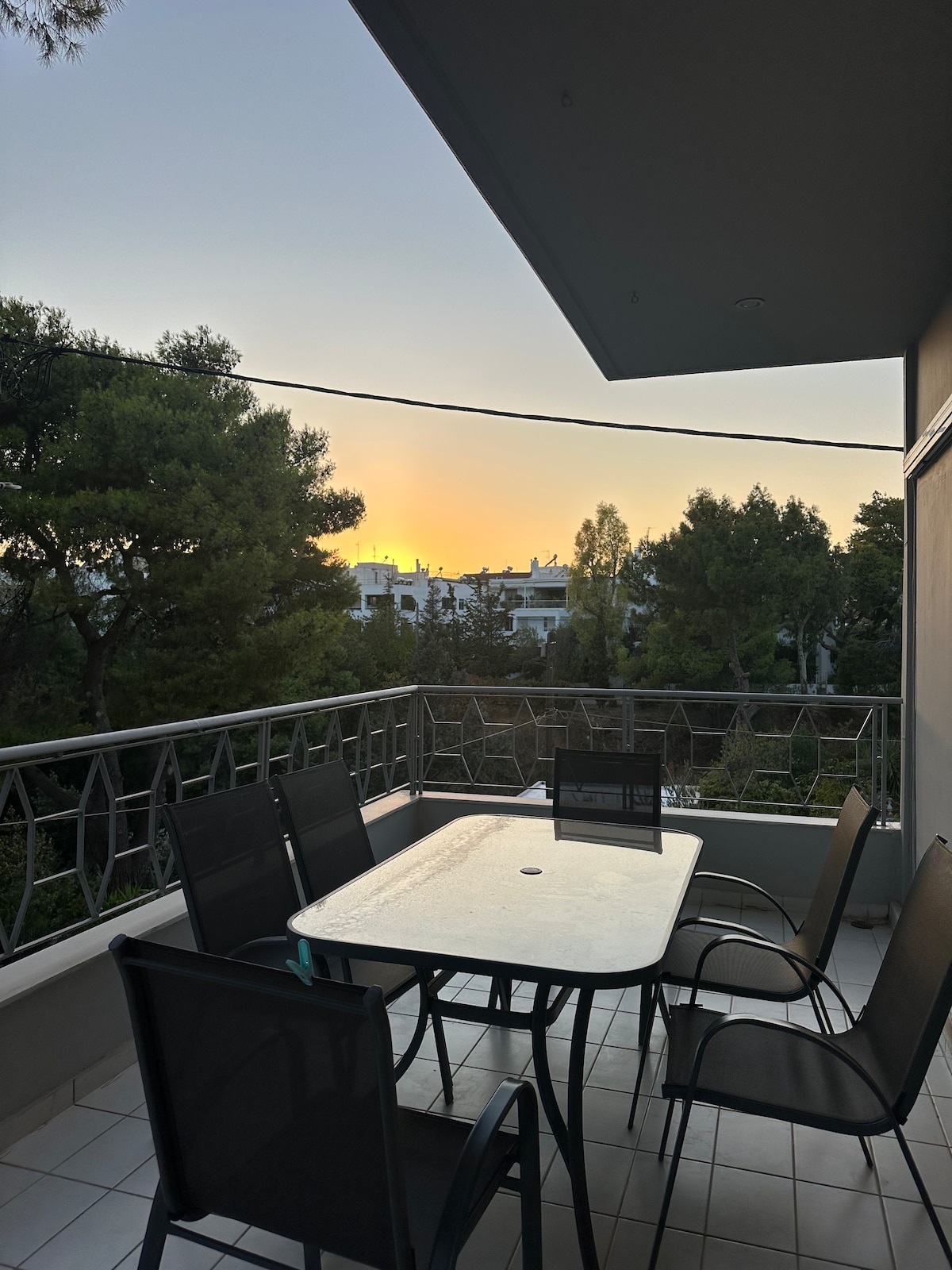 A balcony with a rectangular table surrounded by six chairs is captured during sunset. The warm hues of the sunset are visible in the sky, while lush greenery from surrounding trees enhances the serene atmosphere.