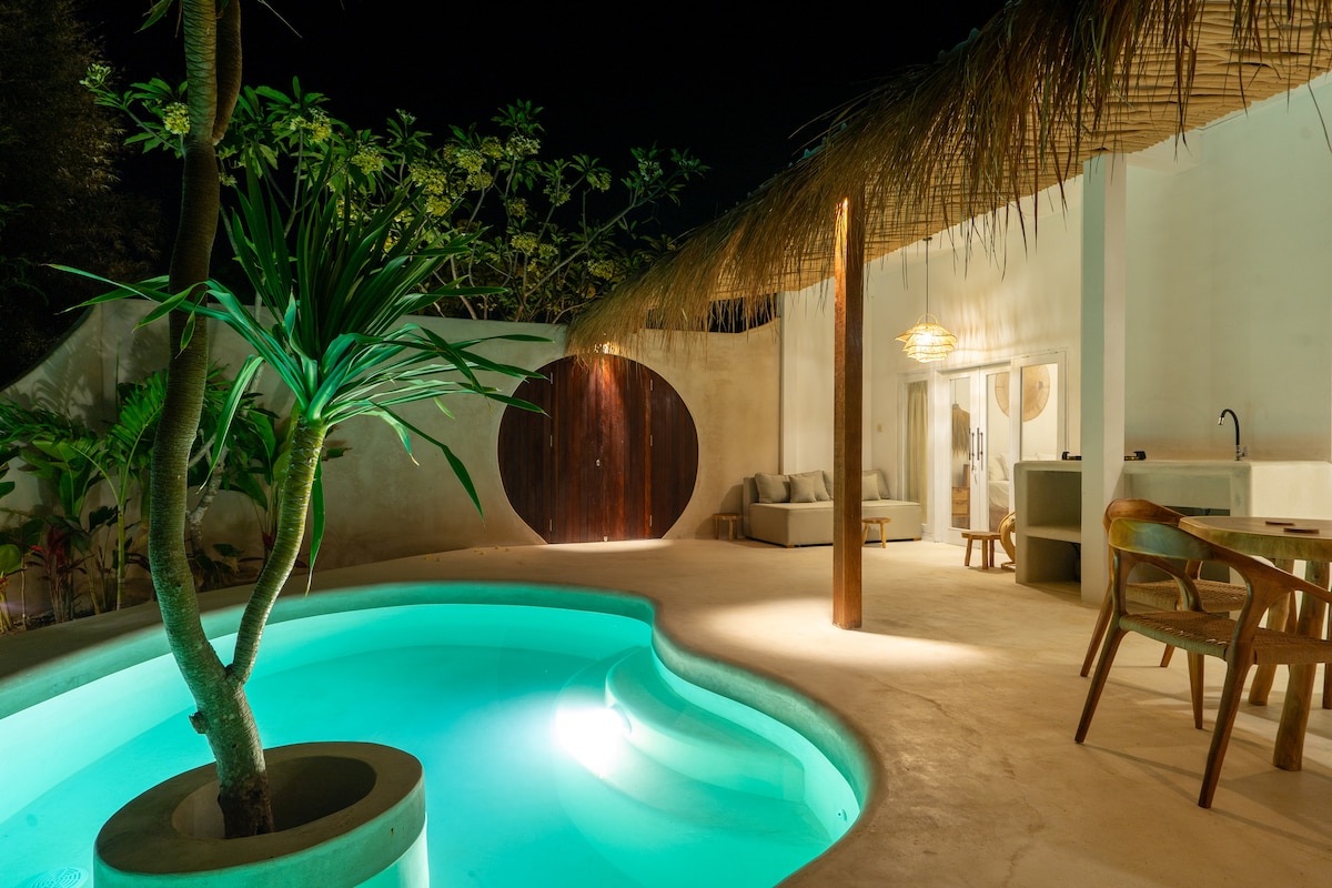 A tranquil pool area is illuminated in soft green hues, surrounded by lush tropical plants. The space features a modern design with a thatched roof overhead and wooden furnishings. A circular wooden door is visible in the background, adding an intriguing architectural element.