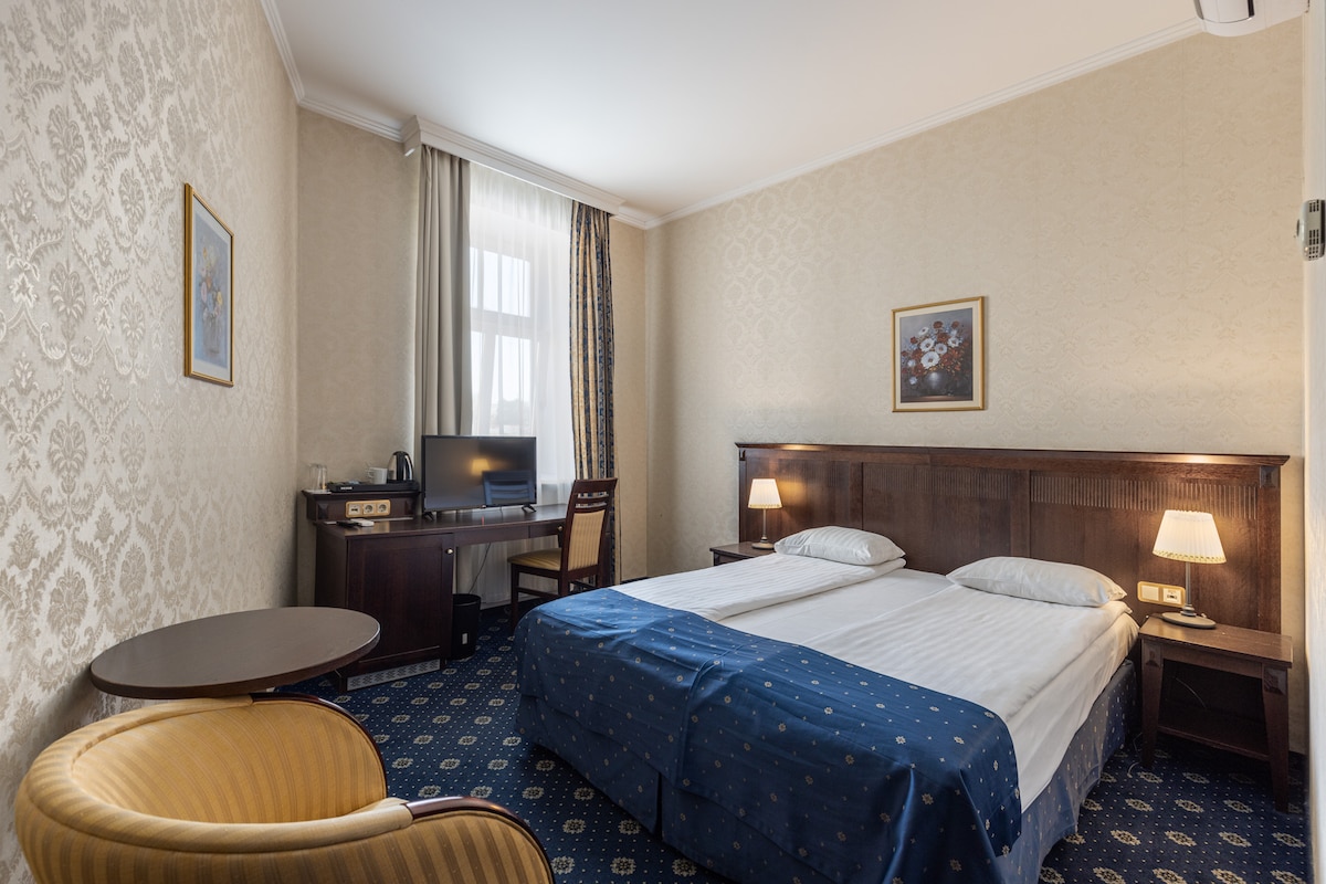 A double hotel room features light-colored wallpaper with a subtle pattern. Two spacious beds are positioned against the wall, accompanied by bedside lamps. A desk with a TV and kettle is present, and a small round table is situated next to a patterned armchair. Natural light enters through a window with plush curtains.