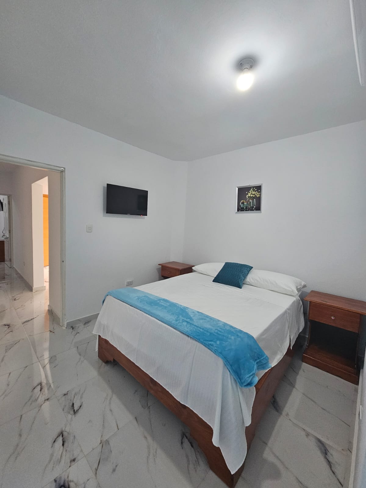 A bright sleeping area is presented with a spacious bed, dressed in white linens and a light blue throw blanket. A small television is mounted on the wall opposite the bed. Light-colored walls and glossy tile flooring contribute to an airy feel.