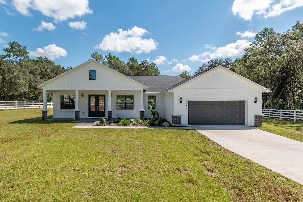 A modern single-story home is nestled on a grassy lawn, surrounded by trees. The house features a gabled roof, large windows, and a covered entryway. A paved driveway leads to a two-car garage, offering access to the welcoming front entrance.