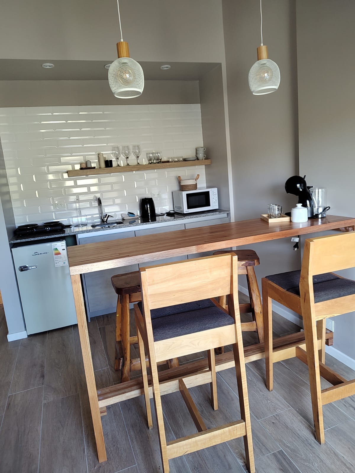 A well-equipped kitchen features modern appliances including a microwave, coffee maker, and electric kettle. The wooden bar with two high chairs offers a casual dining area, while glassware and utensils are neatly arranged on the shelves, complemented by warm lighting from hanging fixtures.