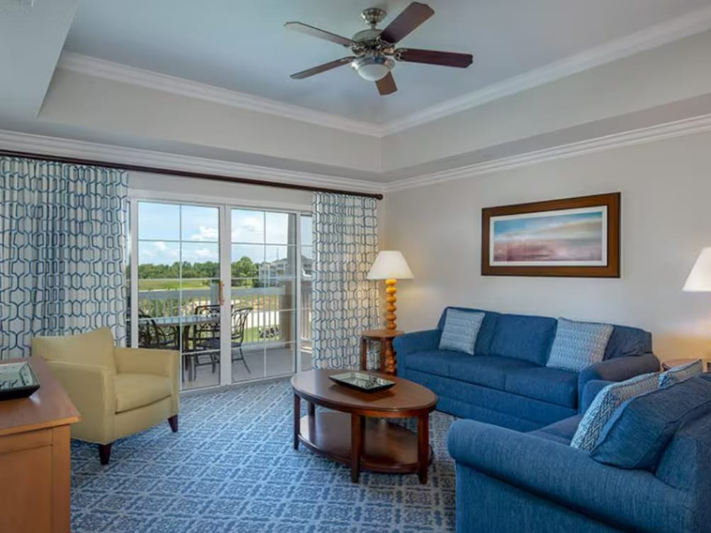A bright and spacious living area features a comfortable blue sofa and matching armchair, complemented by wooden tables. Large glass doors open to a private balcony, inviting natural light and views of the outdoors. A ceiling fan provides a refreshing breeze.
