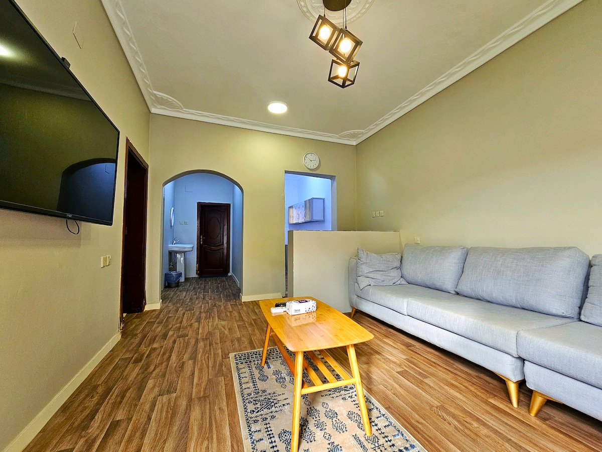 A spacious living area is presented with a light gray sofa positioned in front of a wood coffee table. A large TV is mounted on the wall. An archway leads to a kitchen area, and a clock is visible on the wall.