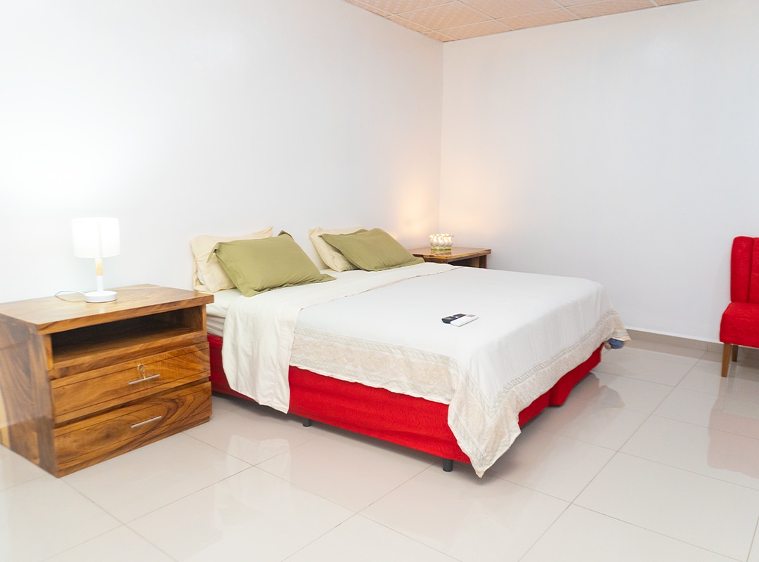 A comfortable room features a spacious bed with light-colored bedding and green pillows. Two bedside tables hold lamps, while a red accent chair is positioned in one corner. The floor is tiled, and soft lighting creates a calm ambiance.