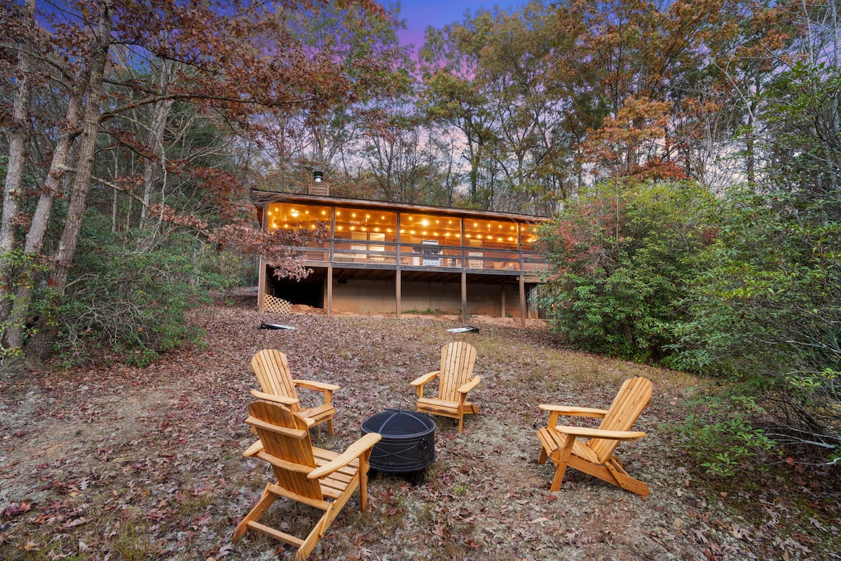 A cozy outdoor seating area contains four wooden Adirondack chairs arranged around a fire pit. String lights illuminate the cabin's expansive deck in the background, surrounded by trees with autumn foliage, providing a serene ambiance for gatherings.