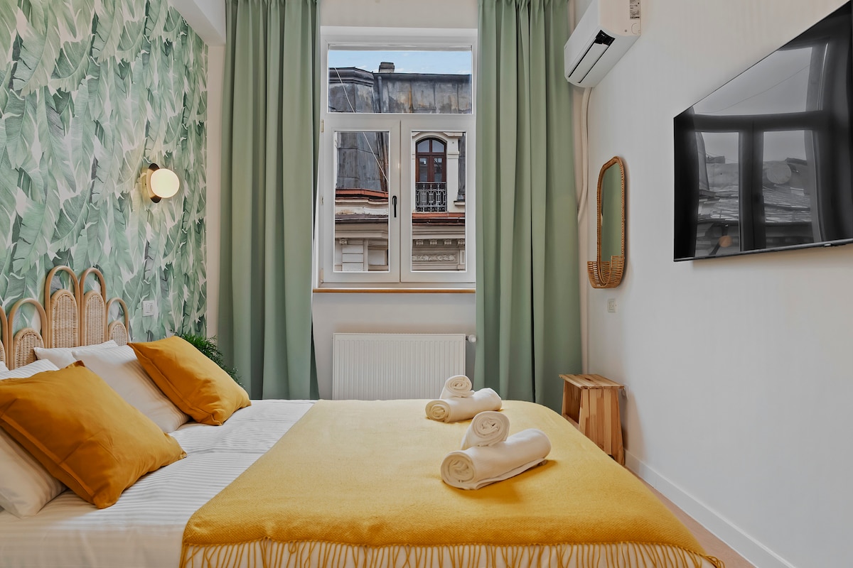 A cozy bedroom features a spacious bed adorned with white linens and mustard yellow accents. Green curtains frame a large window, allowing natural light. A wall decorated with leafy wallpaper adds a touch of nature, while a flat-screen TV and a round mirror are also present.