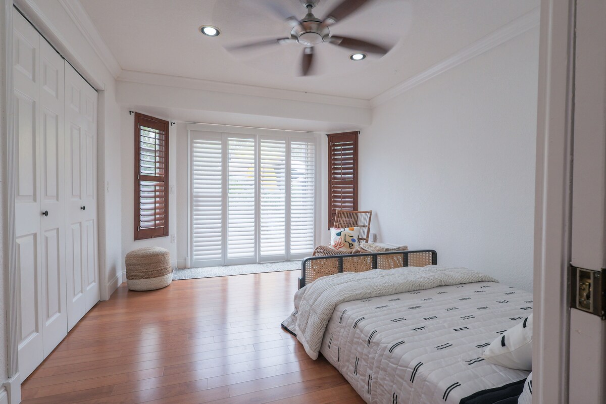 A spacious bedroom is illuminated by natural light streaming through shutters on two sets of glass doors. A comfortable large bed is dressed with a neutral-colored blanket. A ceiling fan adds a cooling feature, while a simple seating area and woven basket provide functional decor.