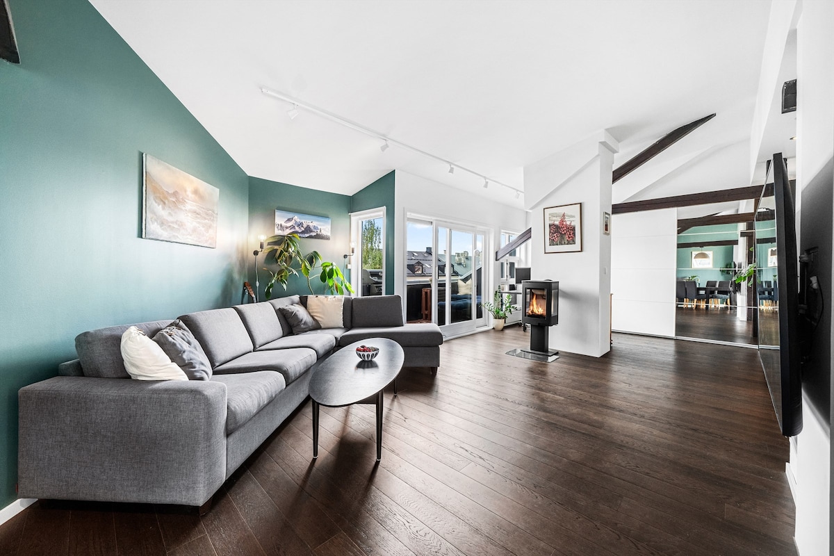 A spacious living area features a gray sectional sofa arranged around a round coffee table, with light hardwood flooring. Large windows provide ample natural light, and a modern fireplace is positioned near the seating area. Decorative plants enhance the inviting ambiance.