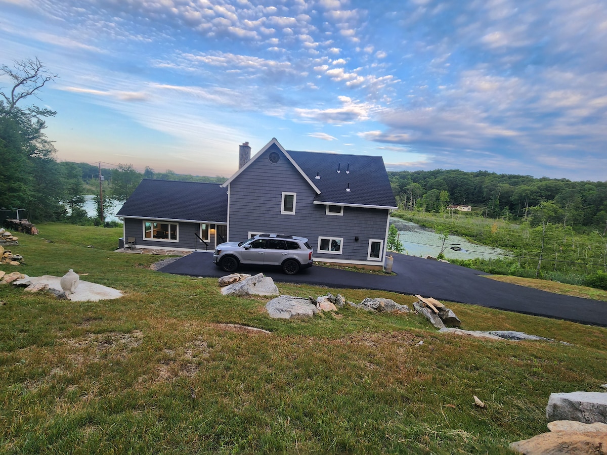 A two-story lake house is depicted, featuring a dark gray siding and a black roof. A vehicle is parked on the paved driveway. Lush green lawn surrounds the house, with natural stone accents visible. A serene lake and green trees are in the background under a blue sky.