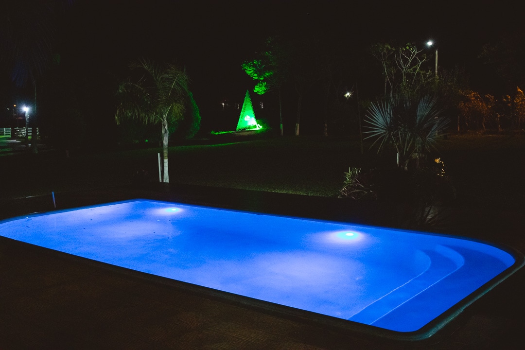 A tranquil pool glows gently under the night sky, featuring vibrant blue lighting and surrounded by lush greenery. Soft illumination from nearby lights creates a serene atmosphere, enhancing the inviting setting for relaxation.