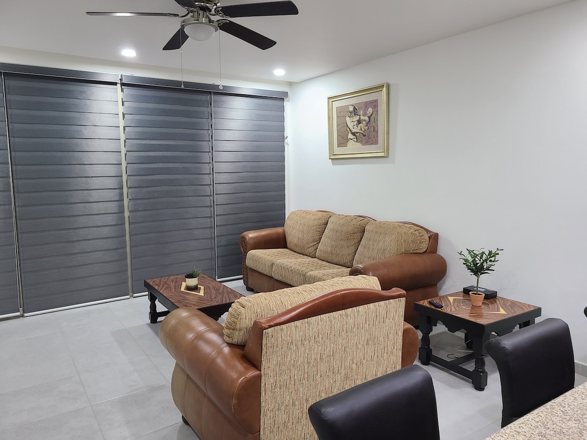 A cozy living area features a mix of light and dark furniture, including a spacious sofa and armchairs. Two wooden coffee tables are positioned in front of the seating. Natural light filters through rolled-up blinds, with a piece of artwork adorning the wall.