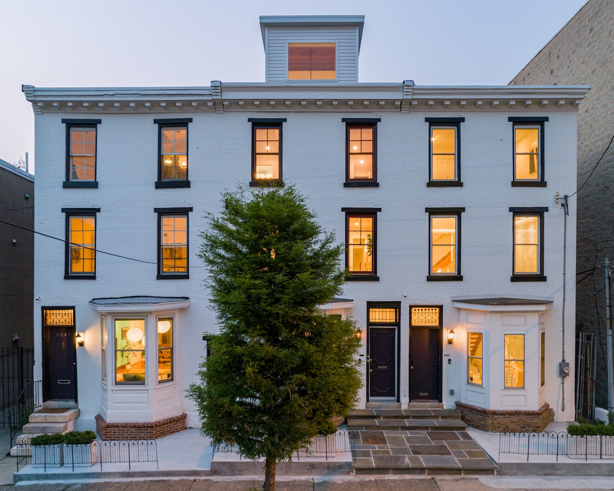 The exterior of a modern townhouse is displayed, featuring a crisp white façade and large, symmetrical windows. A central tree enhances the street view. The three-story structure is complemented by a rooftop feature, with multiple entrance doors and steps leading to the entrance.
