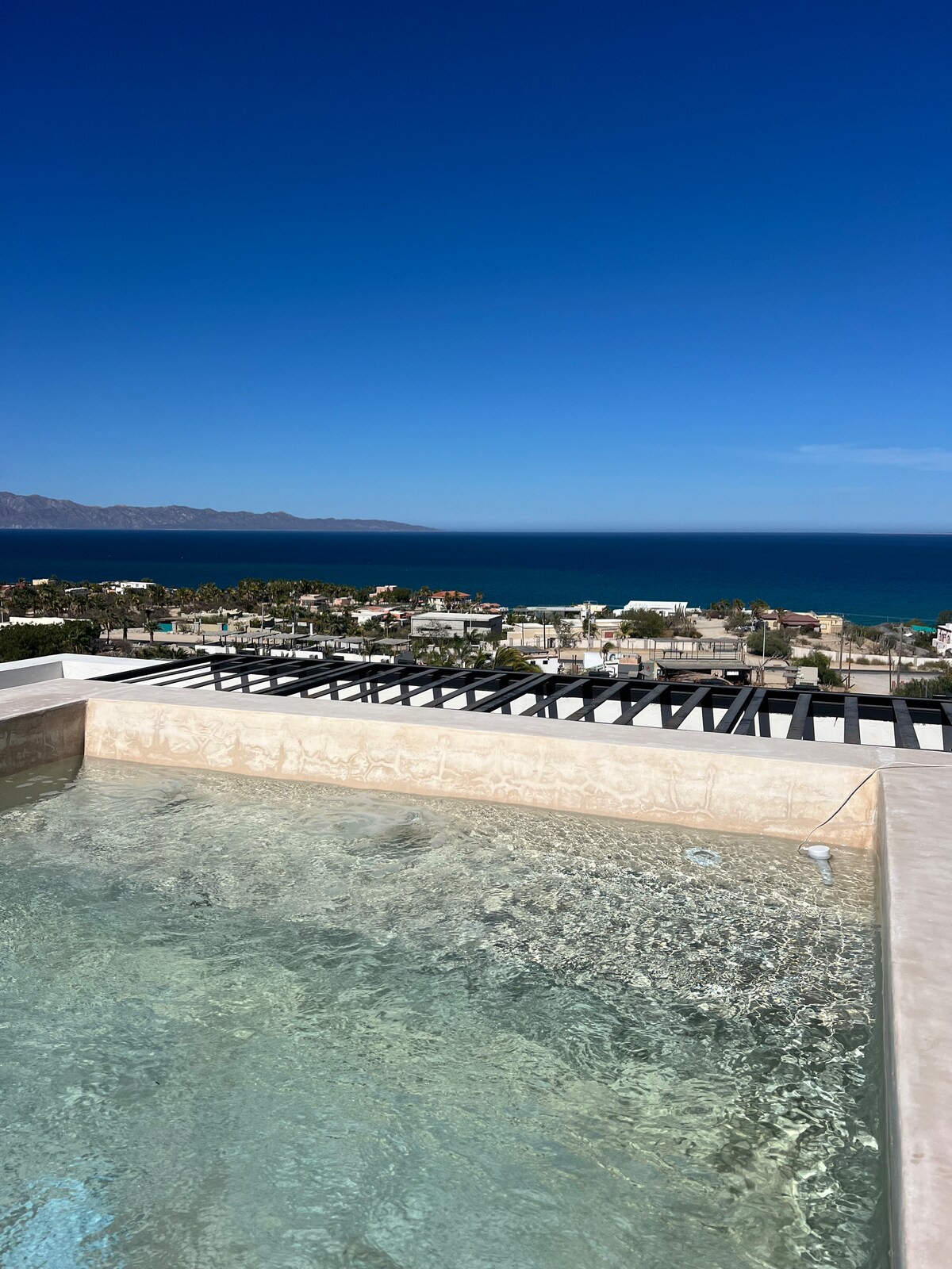 A rooftop area features a hot tub filled with clear water, offering views of the ocean and mountains in the distance. The bright blue sky complements the serene setting, while nearby rooftops and palm trees are visible in the landscape below.