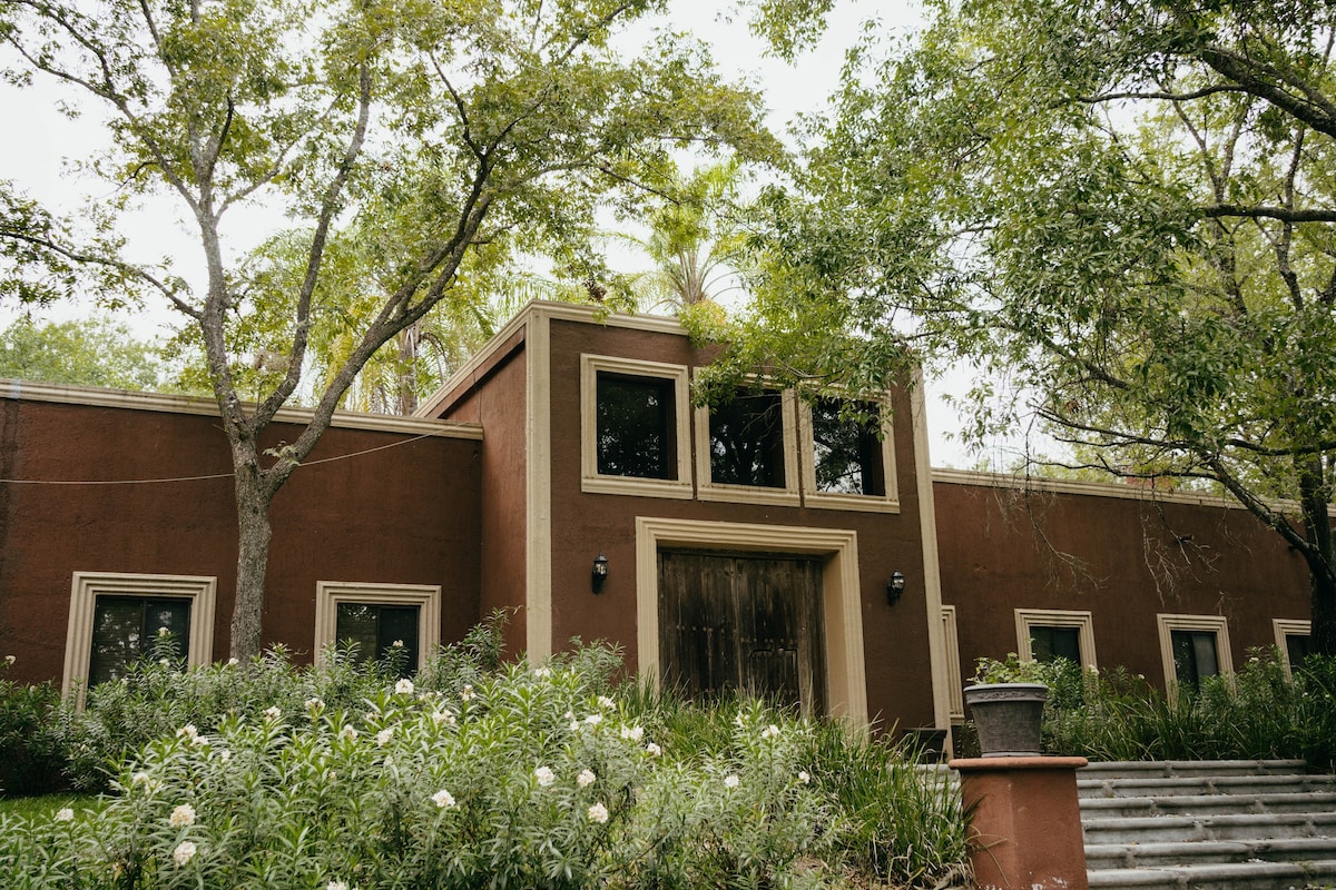 The exterior of a hacienda is framed by lush greenery, featuring a warm terracotta façade and large windows. A set of stone steps leads to the entrance, which is adorned with wooden doors and flanked by well-maintained plants. Tall trees provide natural shade.