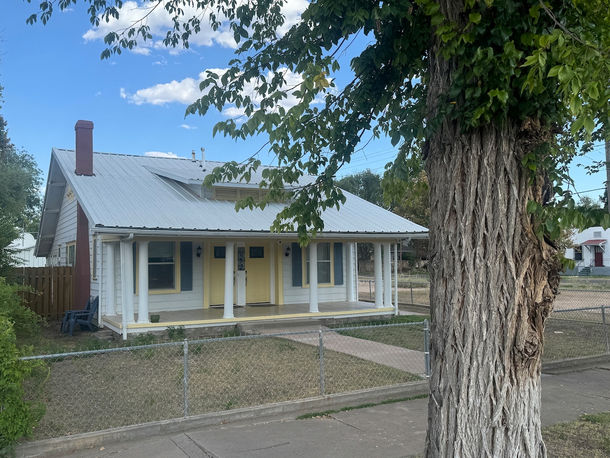 A charming single-story home features a gray metal roof and yellow accents on the front door and shutters. A shaded porch with columns invites relaxation, while a tree and chain-link fence outline the property. An open yard provides space for outdoor activities.