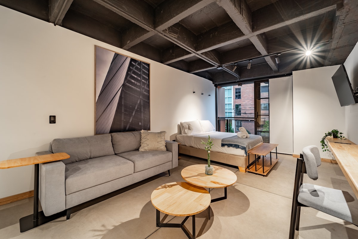 A modern living space features a comfortable sofa and a spacious bed, complemented by minimalist decor. Natural light floods the room through large windows, creating a bright environment. A balcony is accessible, and a desk with two chairs provides a functional workspace.