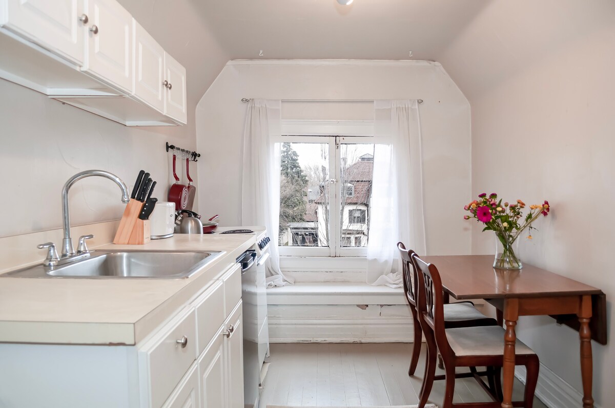 A compact kitchen area features white cabinetry and a double sink. A small wooden dining table with two chairs is positioned by a window adorned with sheer curtains, allowing natural light to filter in. A bouquet of colorful flowers adds a touch of vibrancy.