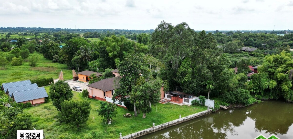 Aerial view showcases the villa and its surrounding landscape, featuring lush greenery and a serene river. The outdoor area includes a spacious lawn, shaded by large trees, with a barbecue area visible near the water's edge, creating a peaceful retreat atmosphere.