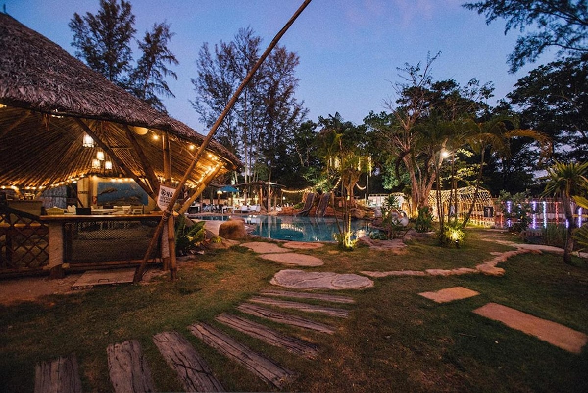 An outdoor area features a beautifully constructed thatched-roof structure, surrounded by greenery and pathways. Soft lights illuminate the space as dusk sets in, highlighting the inviting swimming pool at the center, with lounge chairs positioned nearby for relaxation.