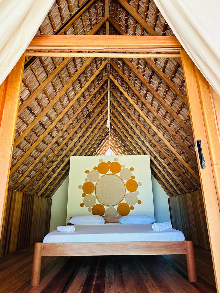 A cozy sleeping area features a natural wood bed with neatly arranged linens. The room's vaulted ceiling is adorned with wood beams, enhancing the spacious feel. A circular wall art piece adds a touch of design, while light enters through the open doorway.