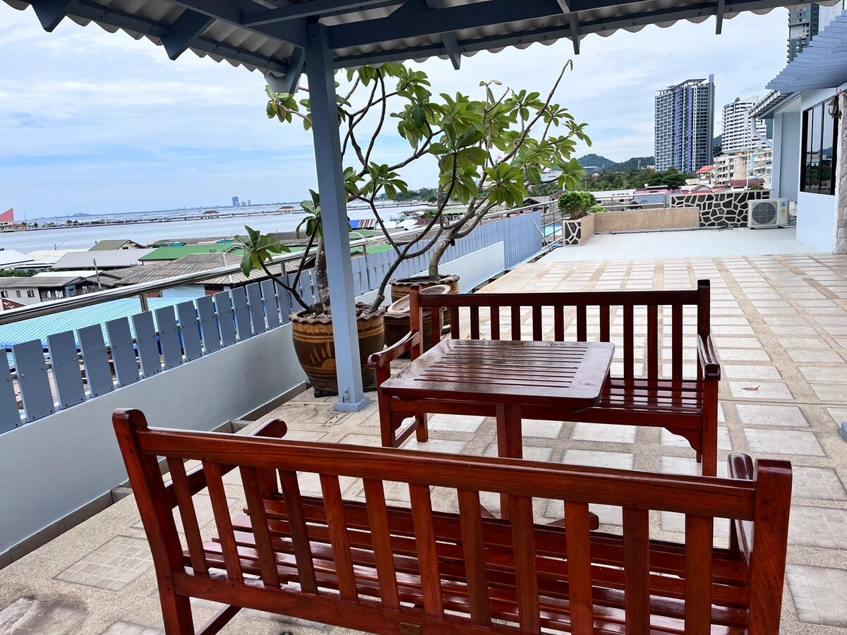 Outdoor seating is arranged on the terrace, featuring wooden benches and a small table. A large potted tree adds greenery, while the view of the sea and nearby buildings can be seen in the background under a cloudy sky.