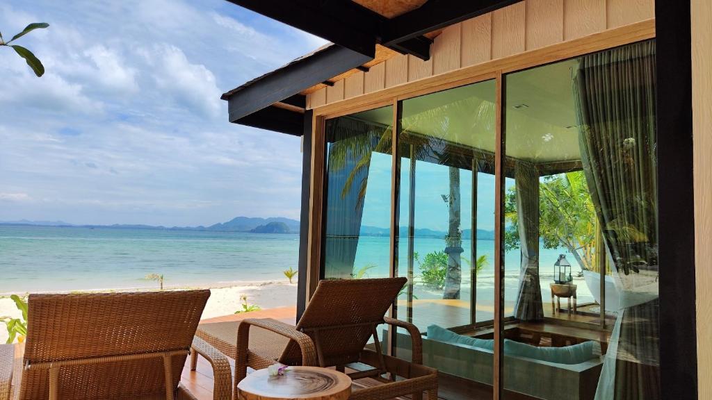 A seating area is positioned under a covered terrace, featuring two wicker chairs and a small table. Large glass doors provide an unobstructed view of the beach and ocean, with palm trees visible in the foreground and distant mountains along the horizon.