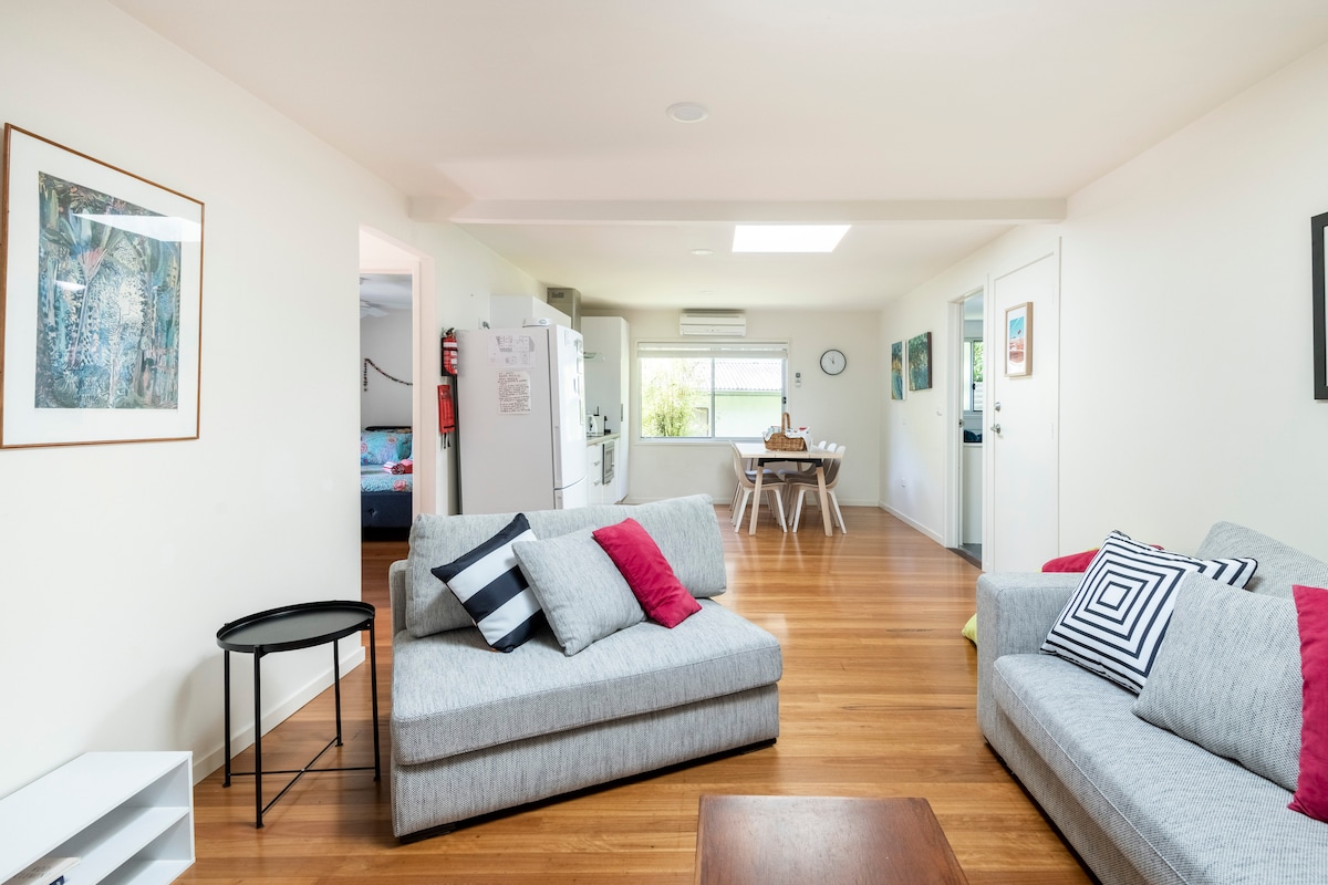 A spacious living area features light wooden flooring and neutral walls. Two grey sofas with colorful cushions are arranged around a wooden coffee table. A dining area with a round table is visible, and a well-equipped kitchen is seen in the background.