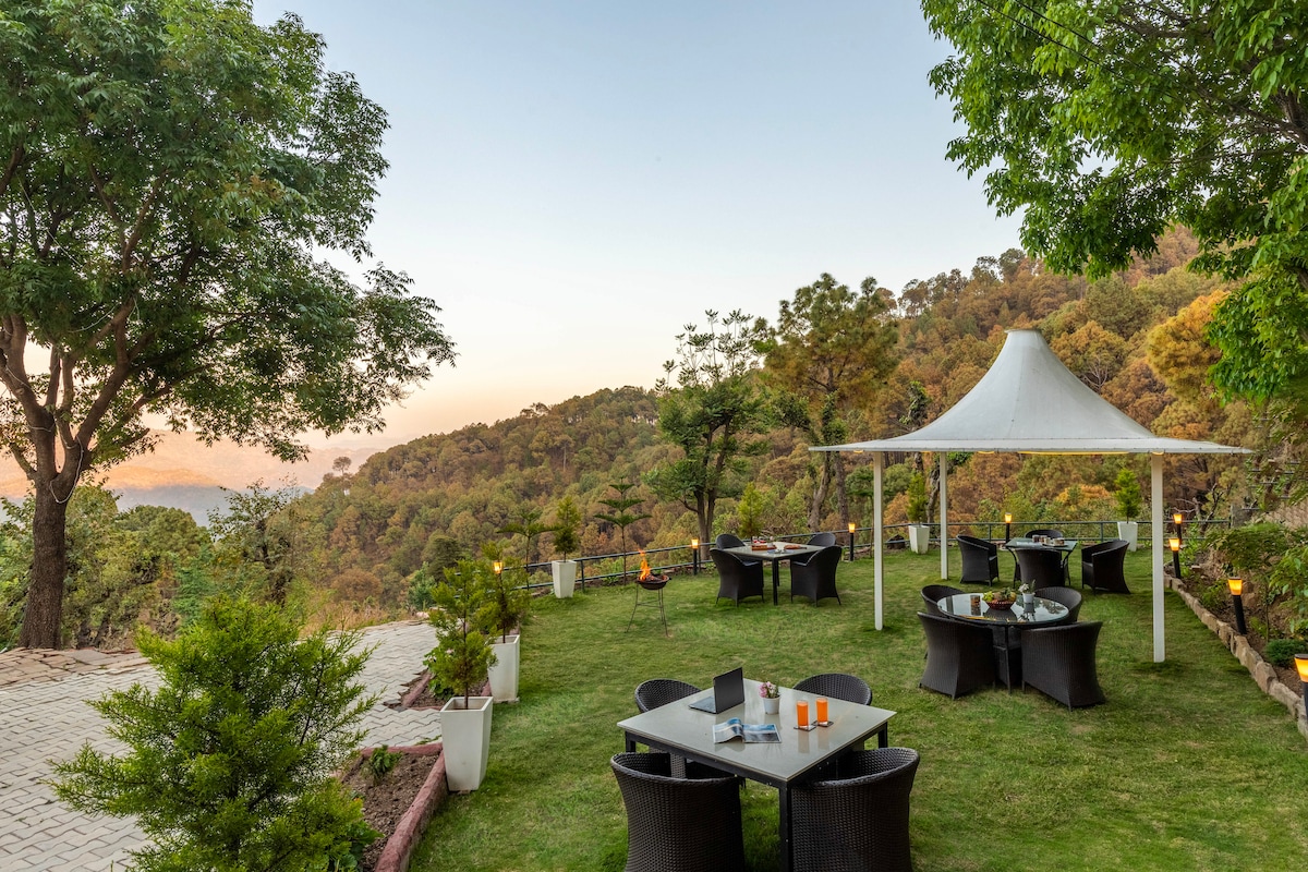 An outdoor dining area is set within a lush garden, featuring tables and chairs arranged under a white canopy. The surrounding greenery offers a scenic backdrop, complemented by distant hill views. Soft lighting enhances the inviting atmosphere of the space.
