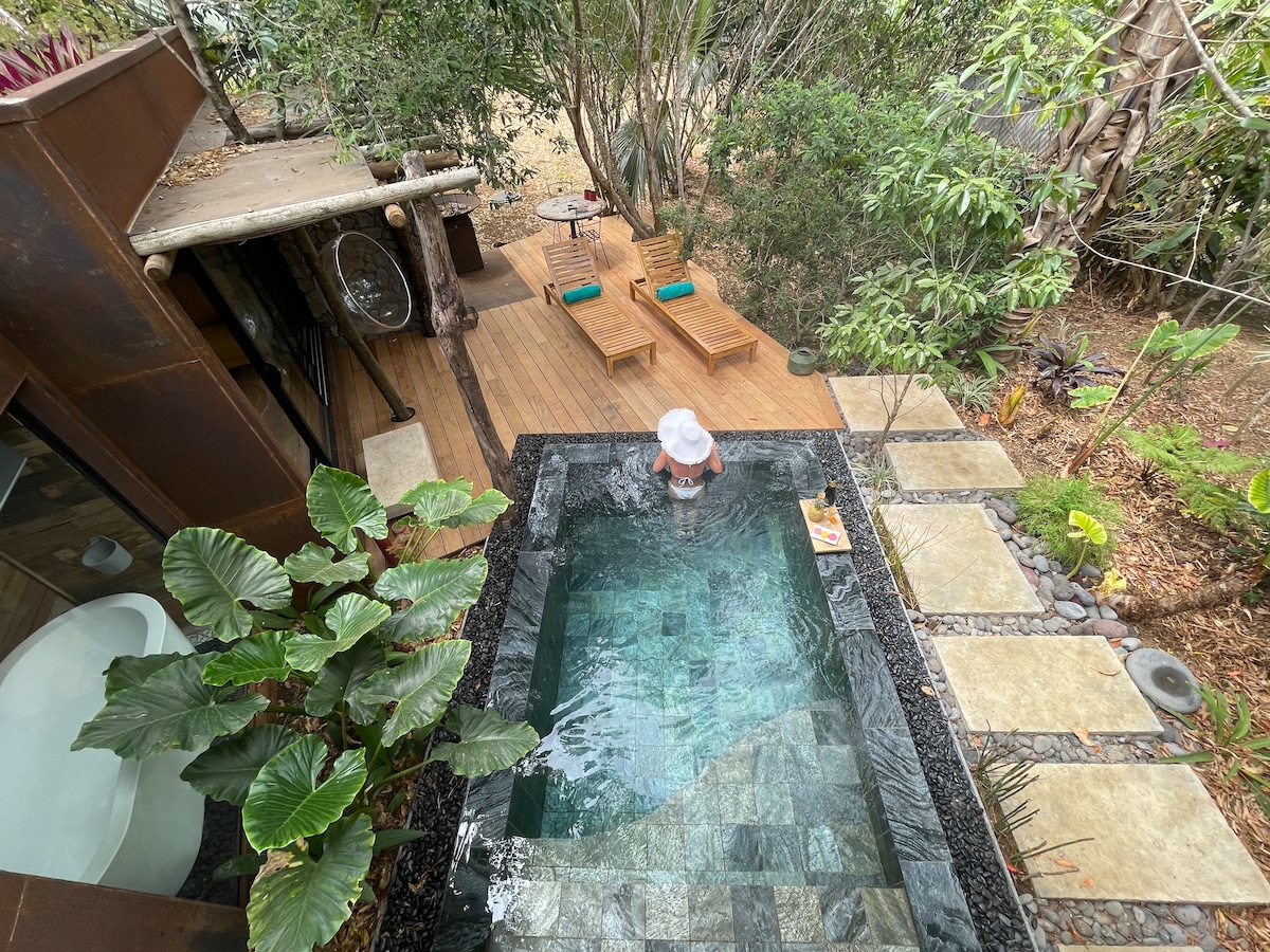 The outdoor area features a private pool nestled amid lush greenery, surrounded by wooden decking. Comfortable lounge chairs are positioned alongside the pool, inviting relaxation. The surrounding natural landscape creates a serene ambiance, enhancing the overall atmosphere of tranquility.