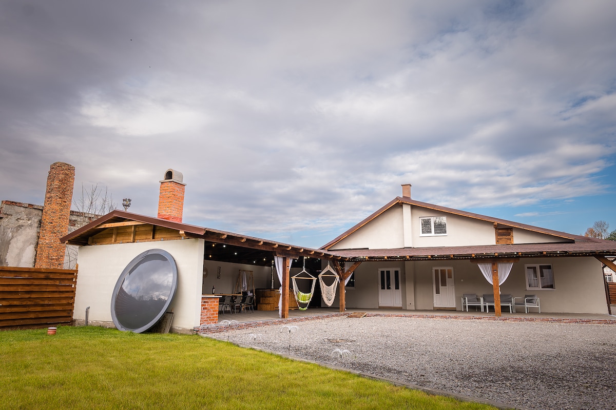 An outdoor space is presented, featuring a large, inviting terrace with hanging hammocks and a round seating area. A well-maintained lawn stretches in front, while the building showcases a combination of rustic and modern architectural elements under a partly cloudy sky.