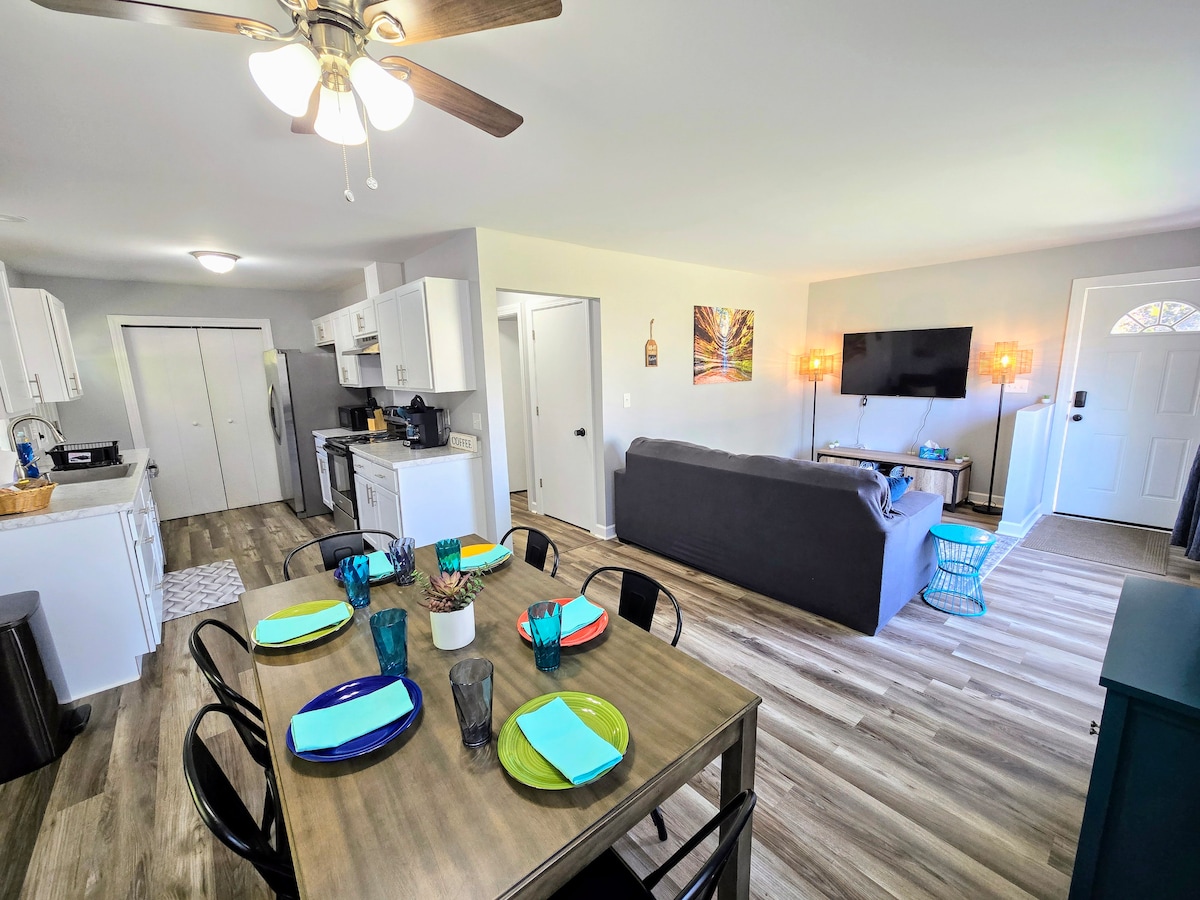 The open living space features a dining table set for six, with colorful plates and glasses. A cozy sofa faces a mounted TV, while a fully equipped kitchen is visible in the background. Natural light enhances the inviting atmosphere through the front door.