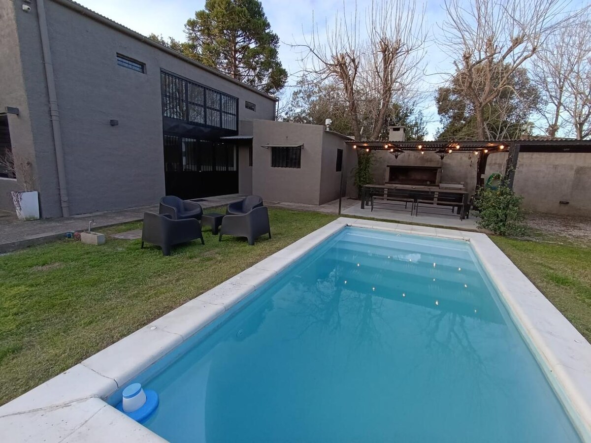 A well-kept outdoor space features a rectangular pool with clear water. Surrounding the pool, grey chairs are positioned on the lawn, providing seating. An open area with string lights above complements a covered outdoor dining area with a grill and table in the background.