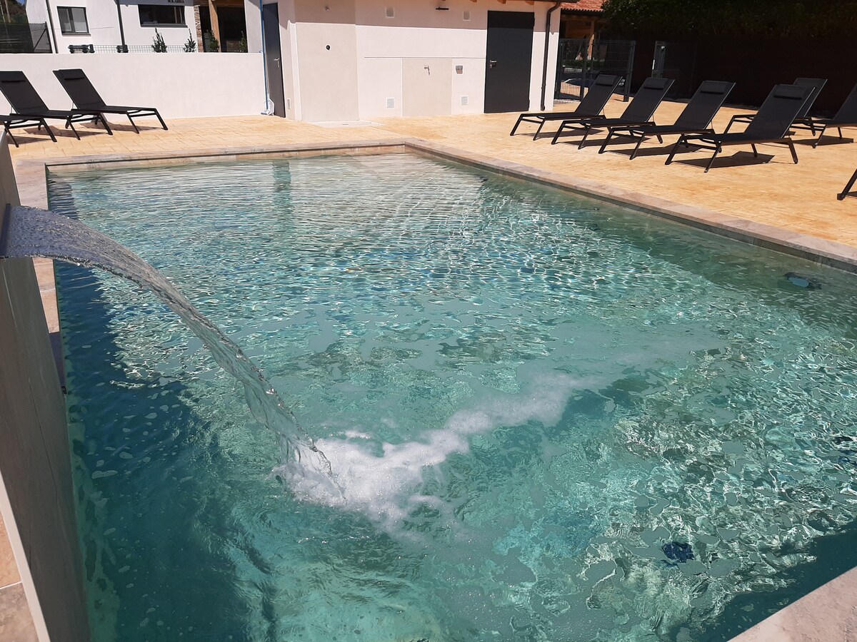 An outdoor pool is surrounded by a sun terrace featuring multiple lounge chairs. Water cascades gently into the pool from a wall-mounted spout, creating a serene atmosphere. The pool's clear water reflects sunlight, inviting relaxation.
