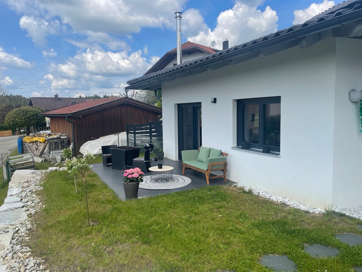 A well-maintained outdoor seating area is set on a grassy patch, featuring modern chairs and a small table. A decorative plant adds color to the space, and the surrounding architecture includes a mix of contemporary and traditional buildings, framed by a bright sky.