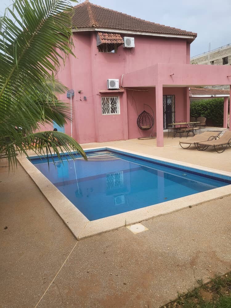 The outdoor area features a clear swimming pool surrounded by a beige paved surface. A small patio is visible, with lounge chairs positioned nearby. The pink exterior of the villa adds a vibrant touch, complemented by green foliage from surrounding plants.