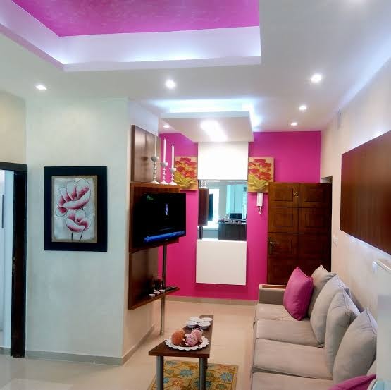 A modern living area is seen, featuring a light sofa with pastel cushions and a decorative table. A vibrant pink wall contrasts with softer shades of the surrounding walls and ceiling. Wall art and mounted lights add to the ambiance. A television is mounted on a wooden panel.