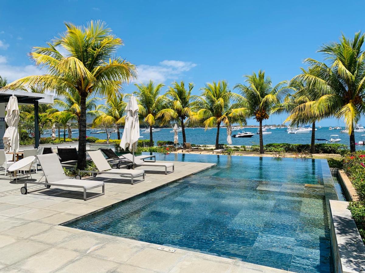 An inviting infinity pool is surrounded by lush palm trees and a scenic view of the ocean. Comfortable lounge chairs are strategically placed for relaxation. The clear blue sky complements the tranquil atmosphere, while boats can be seen gently moored in the distance.