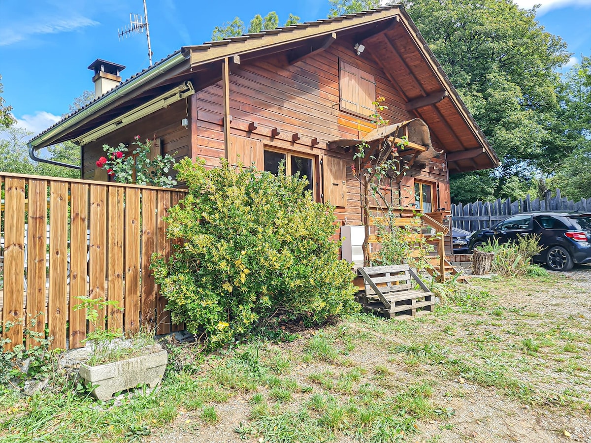 A charming wooden chalet stands surrounded by greenery. The exterior features wooden paneling and a small staircase at the entrance. Flowering plants add a touch of color, while a wooden fence provides privacy. A vehicle is parked nearby on the gravelled area.